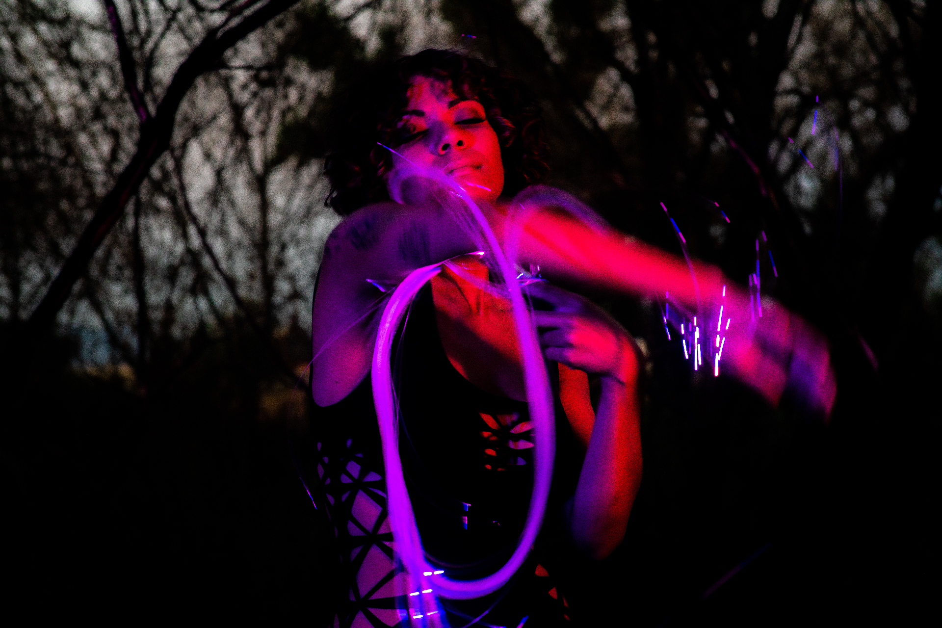 Dancer outdoors at dusk, eyes closed, surrounded by sweeping purple and pink LED light trails